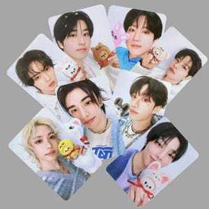 May include: A set of nine photo cards featuring different members of a K-pop group. Each card has a white background and features a close-up of a member holding a small plush toy. The members are all wearing casual clothing and have a variety of hairstyles.
