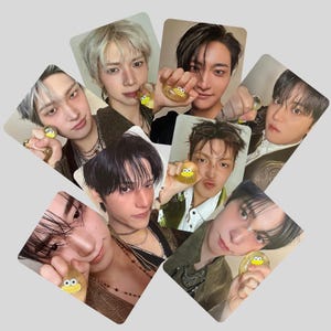 May include: A collection of seven rectangular photo cards featuring portraits of individuals, each holding a small, clear, round, squishy toy with a yellow duck face design. The cards are arranged in a circular fan pattern against a plain background.