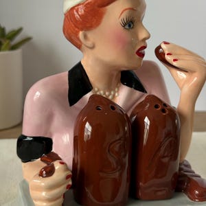 Puede incluir: Un juego de salero y pimentero de cer&aacute;mica con una mujer con gorro de chef y uniforme rosa. El juego incluye dos saleros marrones y una bandeja. La mujer tiene el pelo rojo, l&aacute;piz labial rojo y sostiene un chocolate. Un art&iacute;culo de colecci&oacute;n.