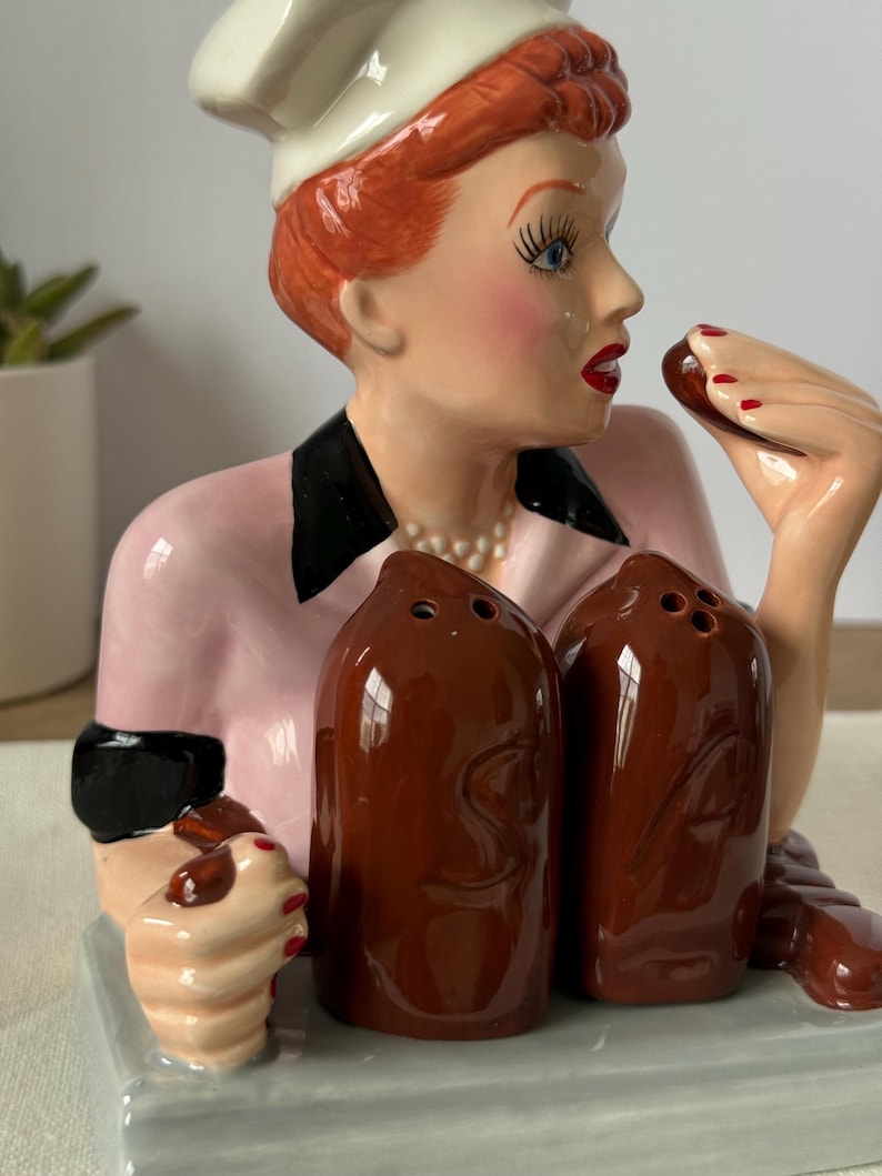 Puede incluir: Un juego de salero y pimentero de cer&aacute;mica con una mujer con gorro de chef y uniforme rosa. El juego incluye dos saleros marrones y una bandeja. La mujer tiene el pelo rojo, l&aacute;piz labial rojo y sostiene un chocolate. Un art&iacute;culo de colecci&oacute;n.