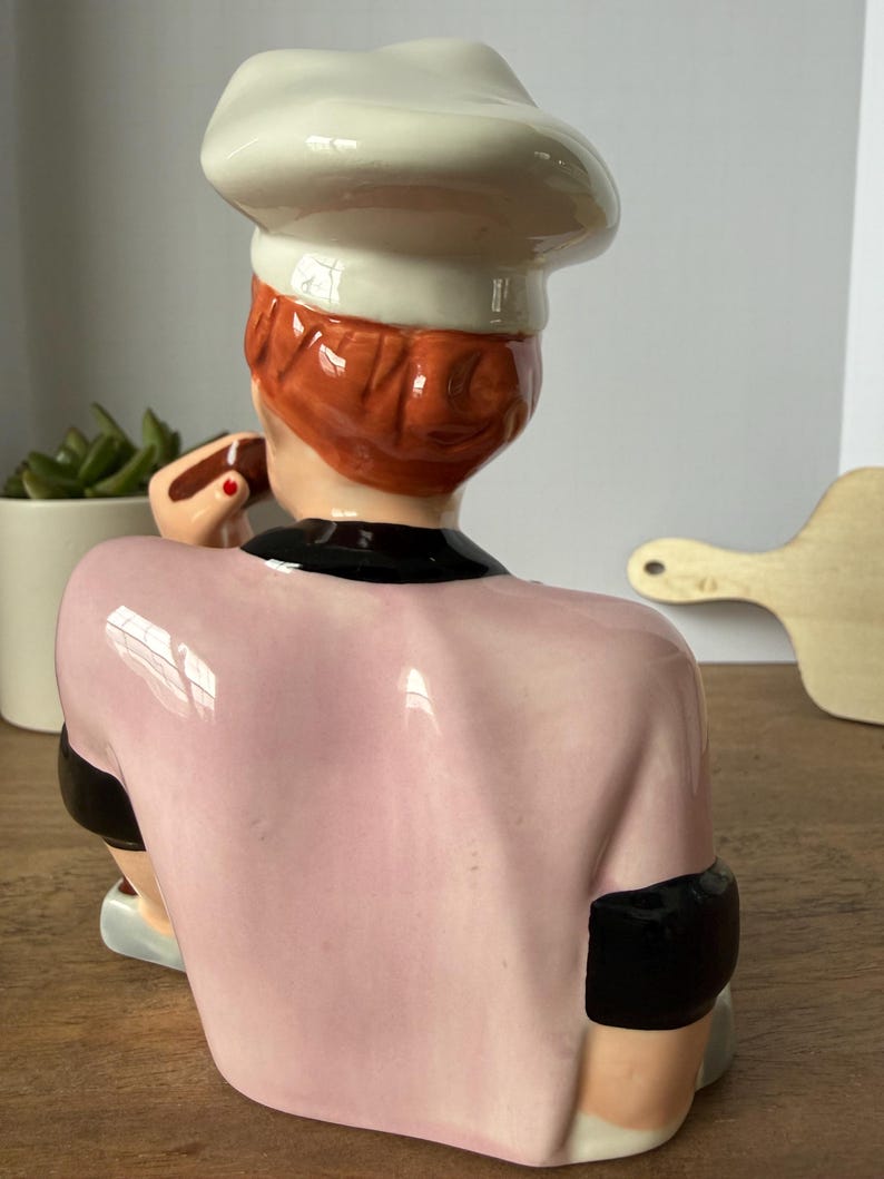 Puede incluir: Figura de cer&aacute;mica de un chef, vista desde atr&aacute;s. El chef lleva un gorro blanco, un uniforme rosa con detalles negros y pelo rojo. La figura sostiene un peque&ntilde;o objeto marr&oacute;n. El fondo incluye una superficie de madera y una peque&ntilde;a planta.