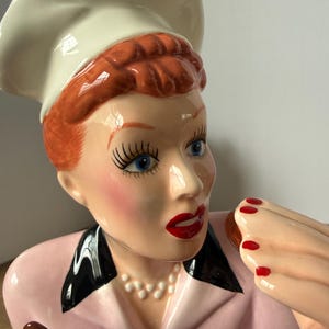 Puede incluir: Figura de cer&aacute;mica de una mujer con gorro de chef y chaqueta rosa, sosteniendo un producto horneado. La figura tiene cabello rojo, ojos azules y l&aacute;piz labial rojo. La mujer lleva un collar de perlas y esmalte de u&ntilde;as rojo.