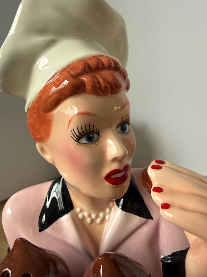Puede incluir: Figura de cer&aacute;mica de una mujer con gorro de chef y chaqueta rosa, sosteniendo un producto horneado. La figura tiene cabello rojo, ojos azules y l&aacute;piz labial rojo. La mujer lleva un collar de perlas y esmalte de u&ntilde;as rojo.