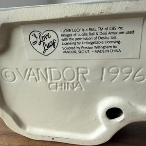 Puede incluir: Base de figura de cer&aacute;mica color crema con el texto "I Love Lucy" y "VANDOR 1996 CHINA" en relieve. La figura tiene un logotipo en forma de coraz&oacute;n y una etiqueta con informaci&oacute;n de licencia.