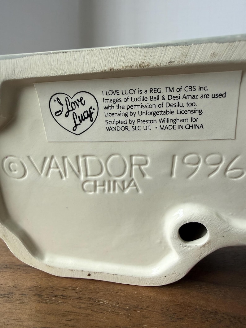 Puede incluir: Base de figura de cer&aacute;mica color crema con el texto "I Love Lucy" y "VANDOR 1996 CHINA" en relieve. La figura tiene un logotipo en forma de coraz&oacute;n y una etiqueta con informaci&oacute;n de licencia.