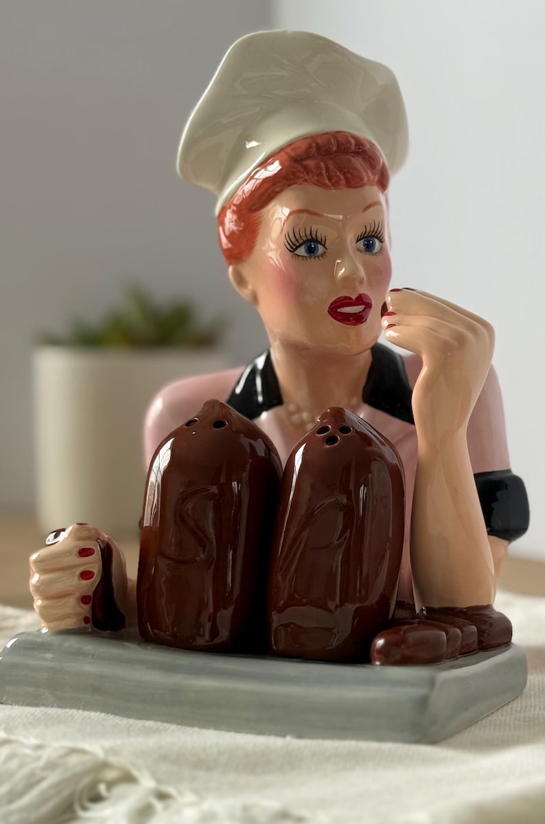 Puede incluir: Un juego de salero y pimentero de cer&aacute;mica con una mujer con gorro de chef y uniforme rosa. Los saleros y pimenteros marrones tienen forma de barril. La mujer tiene el pelo rojo, ojos azules y pintalabios rojo. La base es gris.