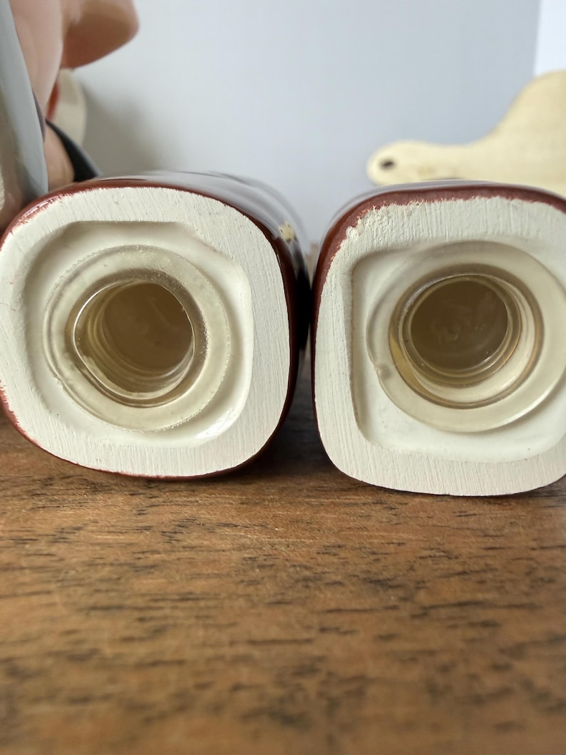 Puede incluir: Primer plano de dos saleros y pimenteros de cer&aacute;mica. Los saleros y pimenteros son de color marr&oacute;n rojizo oscuro con un borde blanco y una inserci&oacute;n de vidrio transparente. Los saleros y pimenteros est&aacute;n sobre una superficie de madera.