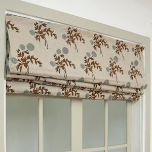 Puede incluir: Una cortina romana beige con un patrón floral repetido en marrón y azul verdoso. La cortina está montada sobre una puerta de vidrio con marco blanco y un patrón de cuadrícula. La tela está doblada, revelando la puerta debajo.