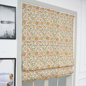 Puede incluir: Una cortina romana con estampado floral en una ventana. La tela presenta un patrón repetido de flores amarillas y naranjas con hojas verdes sobre un fondo crema. El marco de la ventana es blanco y la cortina está parcialmente subida.