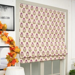 Puede incluir: Una cortina romana con estampado floral en una ventana. La cortina presenta un patrón repetitivo de flores moradas sobre un fondo crema. El marco de la ventana está pintado de color gris claro. Una lámpara blanca con una pantalla plisada está en primer plano.