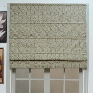 May include: A beige Roman blind with a repeating floral pattern is installed above a white window. The blind is partially lowered, revealing the window's grid pattern. The window is framed by a white trim. Decorative artwork is visible on the wall.