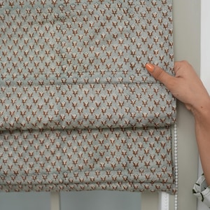 Puede incluir: Una cortina romana con un patrón repetitivo de motivos florales marrones sobre un fondo azul claro. La cortina está parcialmente desplegada, revelando su construcción en capas. Una mano con esmalte de uñas naranja sostiene la cortina.