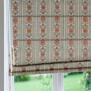 May include: A Roman shade with a repeating floral pattern in peach and gray on an off-white background. The design features vertical stripes with alternating floral and geometric motifs, creating a classic, elegant look. The shade is partially drawn, revealing the window and the beaded pull cord.