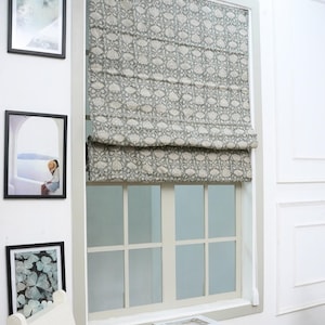 May include: A grey and white floral patterned Roman blind installed on a window with a light grey frame. The fabric features a repeating botanical print with stylised flowers and leaves, partially covering the upper portion of the window pane.