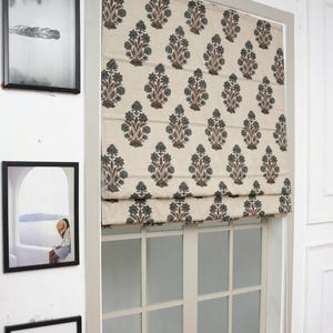 May include: A beige Roman shade featuring a repeating dark green and brown floral block print pattern. The fabric shade is mounted inside a window frame, partially covering the glass panes, with horizontal folds visible across the surface.