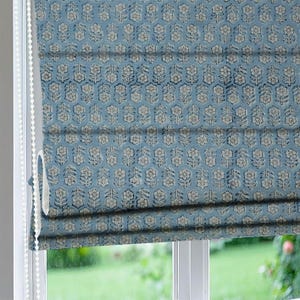 Puede incluir: Una persiana romana azul con un patrón floral repetido en blanco y azul oscuro. La persiana está parcialmente bajada, revelando los pliegues en capas y una vista de la ventana y la vegetación exterior. La persiana es de tela.