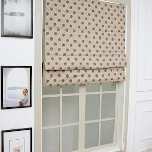 Puede incluir: Una ventana con una persiana romana con estampado beige y gris. La ventana está enmarcada en un color gris claro y tiene un patrón de cuadrícula. Hay cuadros enmarcados en la pared a la izquierda de la ventana. Una pila de libros está en el alféizar.