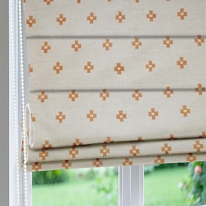 Puede incluir: Estor romano con fondo color crema y un patrón repetido de cruces naranjas. El estor está parcialmente subido, revelando la ventana y la vegetación exterior. Un cordón blanco con cuentas es visible en el lado izquierdo.