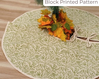 May include: A round, cream-colored Christmas tree skirt with a green floral block print pattern. The skirt has a cream-colored border and ties. The text "Block Printed Pattern" is visible. A bundle of fall leaves rests on the skirt.
