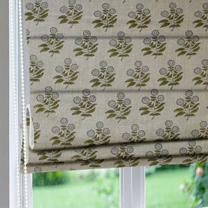 Puede incluir: Una cortina romana con un patrón floral repetido. El diseño presenta flores de color gris claro con tallos y hojas verdes sobre un fondo beige. La cortina está colgada frente a una ventana.