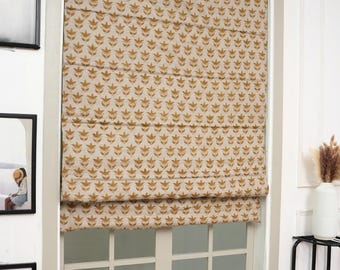 May include: A Roman shade covering a window, featuring a repeating pattern of small, golden-brown floral motifs on a light beige fabric background. The shade is partially raised, creating horizontal folds across the surface of the window covering.