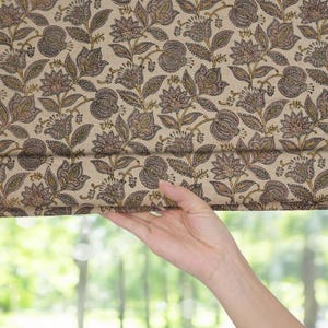 Puede incluir: Una tela estampada floral con un fondo beige y un diseño repetido de flores estilizadas en tonos de marrón, verde y morado. La tela está siendo sostenida por una mano, revelando un fondo verde borroso.