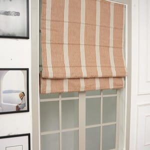 Puede incluir: Una persiana romana a rayas beige y rojas está instalada dentro de un marco de ventana blanco. La persiana está parcialmente bajada, revelando el patrón de cuadrícula de la ventana. La ventana está rodeada de obras de arte enmarcadas y una cesta de frutas.