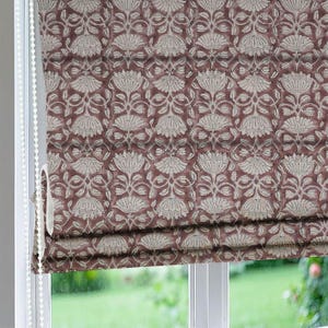 Puede incluir: Una cortina romana con un patrón floral repetido en crema y burdeos. La tela es visible en una ventana, con un cordón de cuentas blanco en el lado izquierdo. El diseño floral presenta flores y hojas estilizadas.