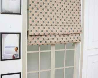 May include: A roman shade featuring a beige fabric base with a repeating pattern of dark teal circular dots surrounded by small orange stippled rings. The shade is mounted inside a window frame with a grid of white muntins and a light beige trim.