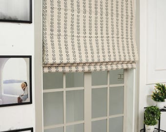 May include: A Roman shade featuring a cream-colored fabric with vertical rows of dark gray leaf-like patterns. The shade is mounted inside a window frame, partially covering the upper portion of a window with a white grid-style pane design.
