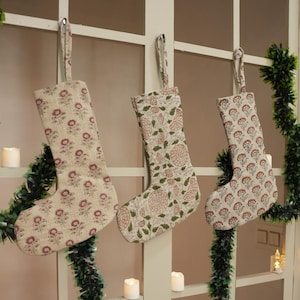 May include: Three Christmas stockings with floral patterns hang from a window. The stockings have a beige background with red and green floral designs. A green garland and candles decorate the window.