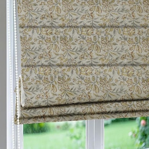 May include: A Roman shade window covering featuring a repeating floral pattern in shades of muted yellow, brown, and grey on a light cream background. The fabric is folded into horizontal pleats with a white beaded pull chain mechanism on the side.