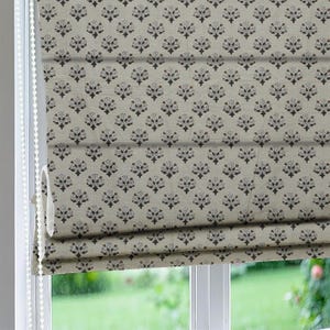 Puede incluir: Una persiana romana beige con un patrón repetido de motivos florales estilizados en marrón oscuro, blanco y burdeos. La persiana está parcialmente enrollada, revelando su construcción en capas y un cordón de cuentas.