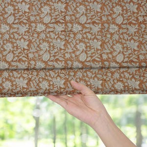 Puede incluir: Primer plano de una tela marrón y crema con estampado floral. La tela presenta un diseño repetido de flores y hojas. La tela está sujeta por una mano, con un fondo verde borroso.