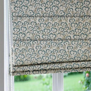 Puede incluir: Una cortina romana con un patrón floral repetido en verde azulado y beige sobre un fondo blanquecino. La cortina está parcialmente bajada, revelando la ventana y una vista del exterior. La cortina es de tela y tiene un cordón.