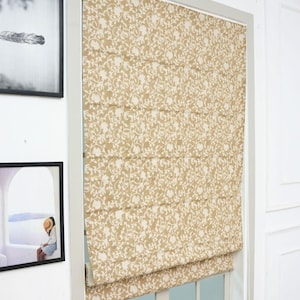 May include: A tan roman shade featuring a repeating white floral and vine pattern. The fabric shade is mounted inside a window frame and is partially raised, creating horizontal folds across the surface of the window covering.