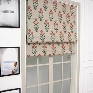 Peut inclure: Un store romain beige à motifs floraux est présenté dans un cadre de fenêtre blanc. Le motif présente des fleurs rouges répétées avec des tiges vertes. Des œuvres d'art encadrées entourent la fenêtre.