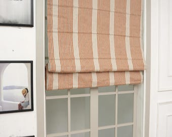 May include: A Roman shade window covering featuring a vertical striped pattern in burnt orange and off-white. The fabric shade is partially raised, creating a horizontal fold at the bottom, and is mounted inside a white window frame.