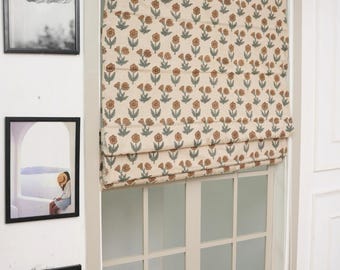 May include: A Roman shade window covering featuring a repeating floral pattern in muted brown and sage green on a cream-colored fabric. The shade is partially raised, displaying horizontal folds, and is mounted inside a white window frame.