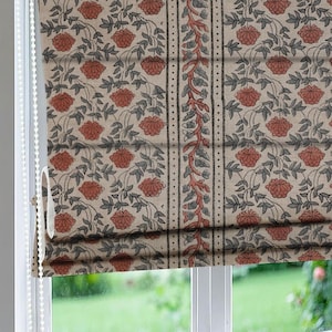 Puede incluir: Una persiana con estampado floral. La tela presenta un patrón repetitivo de flores rojas y hojas grises sobre un fondo beige. Una franja vertical con un diseño de vid y líneas punteadas añade detalles. La persiana está parcialmente enrollada, revelando la ventana.