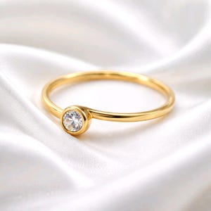 Könnte beinhalten: Ein dünner goldener Ring mit einem einzelnen runden, klaren, facettierten Stein in einer Zargenfassung. Die Ringschiene ist glatt und poliert und bildet eine durchgehende Kreisform, die den Glanz und die Klarheit des Steins betont.