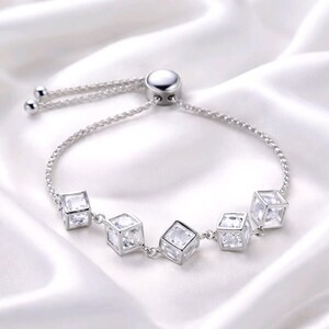 May include: A silver bracelet featuring five clear cubic charms. The bracelet has a fine chain and an adjustable slider clasp. The cubes appear to be filled with clear stones, reflecting light. The bracelet is displayed on white, silky fabric.