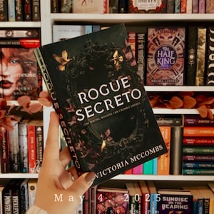 May include: A close-up of a book titled "Rogue Secreto" by Victoria McCombs. The book cover features a dark background with gold accents and the text "One wrong whisper can crumble empires".