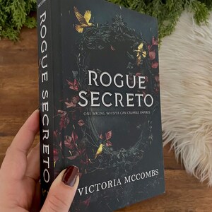 May include: A hardcover book titled "Rogue Secreto" by Victoria McCombs. The cover is dark with gold and red accents, featuring a floral design and a bird. The spine reads "Rogue Secreto" vertically. The book is held by a person.