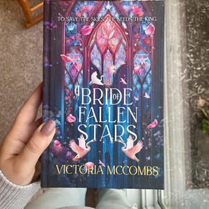 May include: A book cover for "A Bride of Fallen Stars" by Victoria McCombs. The cover features a stained-glass design with pink and blue hues, two pink birds, and falling petals. The text "TO SAVE THE SKIES SHE NEEDS THE KING" is at the top.