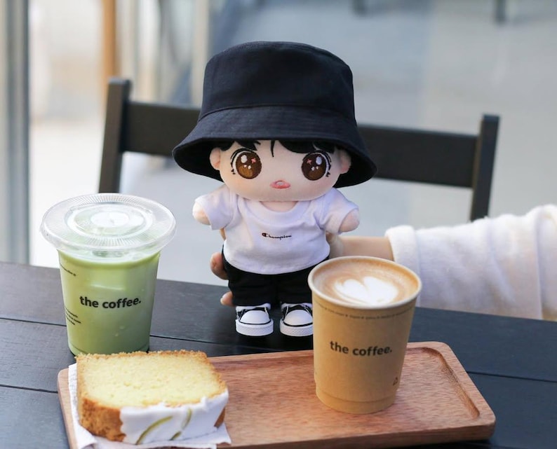 May include: A plush doll wearing a black bucket hat, white t-shirt, black pants, and white sneakers. The doll is holding a cup of coffee and is sitting at a table with a matcha drink, cake, and a coffee cup. The coffee cup and matcha drink have the text "the coffee."