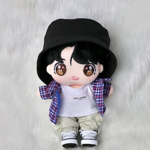 May include: A plush doll wearing a black bucket hat, a white t-shirt with "SAINT LAURENT PARIS" printed on it, and a red, white, and blue plaid shirt. The doll is also wearing khaki pants and white sneakers.