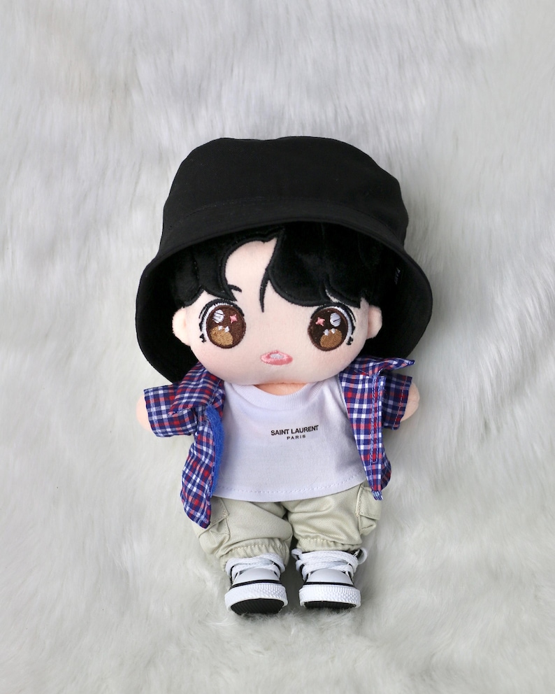 May include: A plush doll wearing a black bucket hat, a white t-shirt with "SAINT LAURENT PARIS" printed on it, and a red, white, and blue plaid shirt. The doll is also wearing khaki pants and white sneakers.
