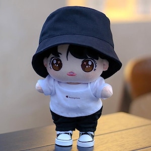 May include: A plush doll wearing a black bucket hat, a white t-shirt with "Champion" printed on it, black shorts, and white sneakers with black accents. The doll has large, expressive eyes and a neutral expression.