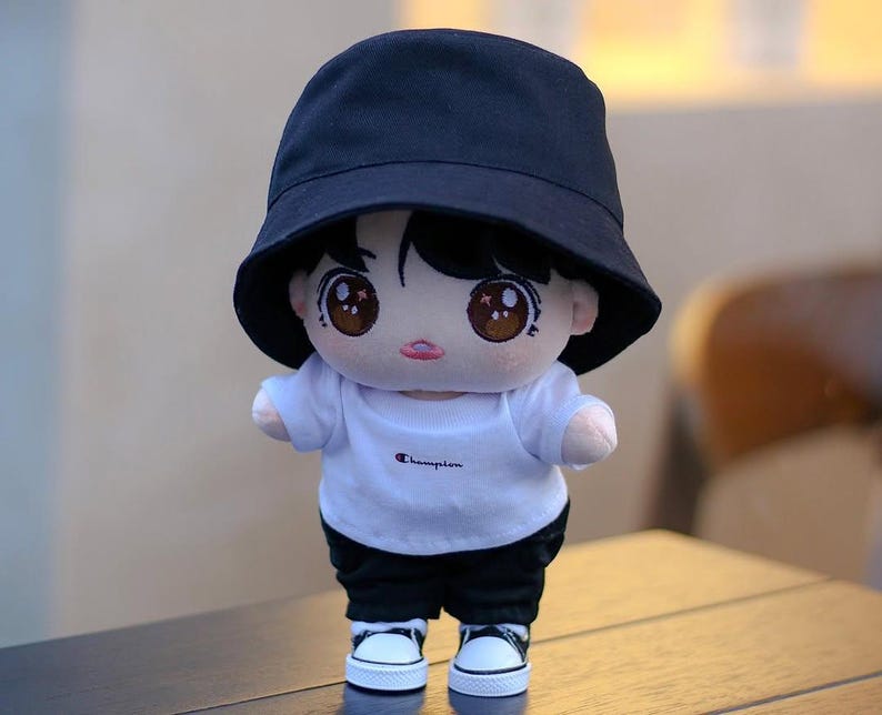 May include: A plush doll wearing a black bucket hat, a white t-shirt with "Champion" printed on it, black shorts, and white sneakers with black accents. The doll has large, expressive eyes and a neutral expression.